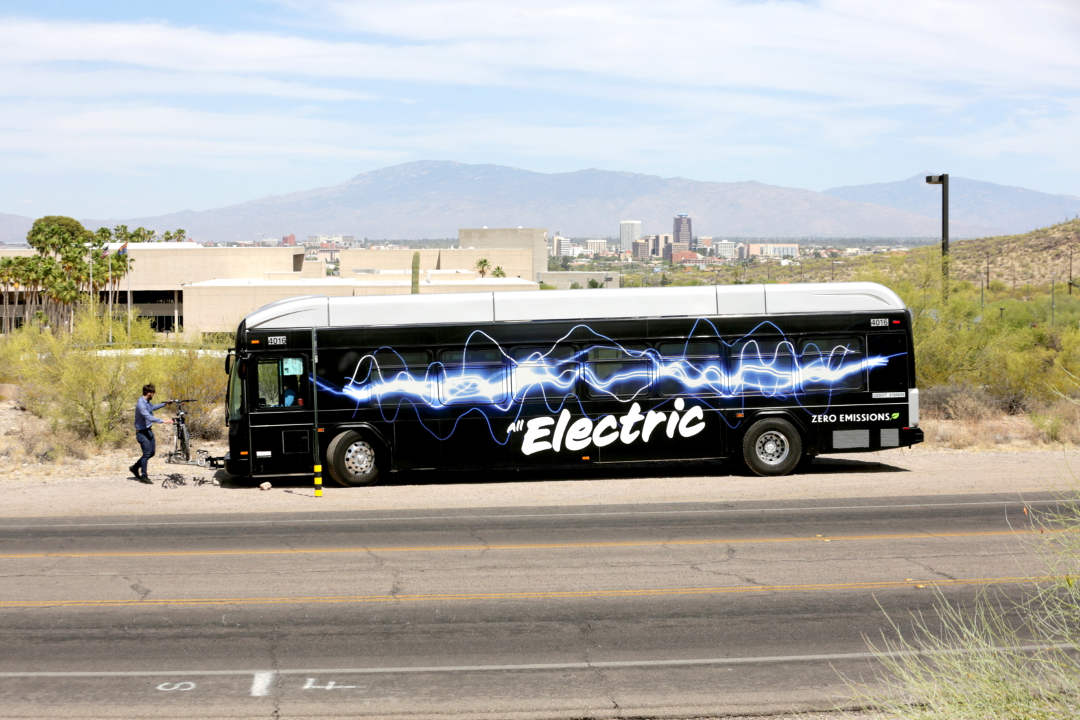 A Sun Tran electric bus drops off a passenger with the Tucson city skyline is visible in the background.