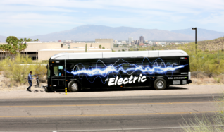 A Sun Tran electric bus drops off a passenger with the Tucson city skyline is visible in the background.